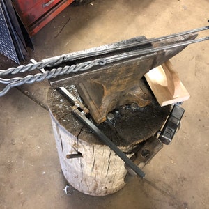 May include: A metal blacksmithing anvil with a twisted metal rod resting on top. The anvil is sitting on a wooden stump.