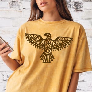 May include: Mustard yellow t-shirt featuring a brown geometric eagle design. The short-sleeved shirt has a crew neck and a slightly oversized fit. The eagle design is centered on the front of the shirt.