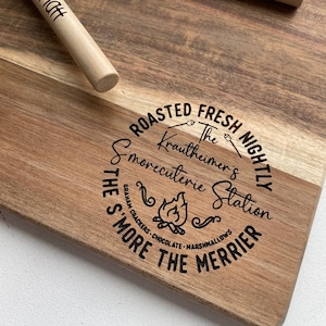 May include: A wooden chopping board with a burnt-in design that says "Roasted Fresh Nightly The Krautheimer's S'morecuterie Station The S'more The Merrier Graham Crackers Chocolate Marshmallows". The board has two wooden skewers with red handles and the names "Emma" and "Matt" engraved on them.