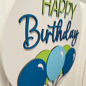 K&ouml;nnte beinhalten: Ein rundes, wei&szlig;es Holzschild mit den Worten "HAPPY Birthday" in Gr&uuml;n und Blau. Am unteren Rand des Schildes sind drei Ballons in Blau- und Gr&uuml;nt&ouml;nen befestigt. Das Schild ist ein dekorativer Artikel f&uuml;r eine Geburtstagsfeier.