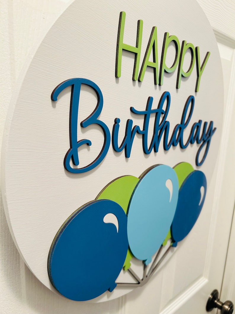 K&ouml;nnte beinhalten: Ein rundes, wei&szlig;es Holzschild mit den Worten "HAPPY Birthday" in Gr&uuml;n und Blau. Am unteren Rand des Schildes sind drei Ballons in Blau- und Gr&uuml;nt&ouml;nen befestigt. Das Schild ist ein dekorativer Artikel f&uuml;r eine Geburtstagsfeier.