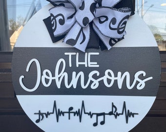 May include: A white and black round wooden door hanger with the text "THE Johnsons" and a musical note heartbeat design. A black and white ribbon with musical notes is tied at the top.