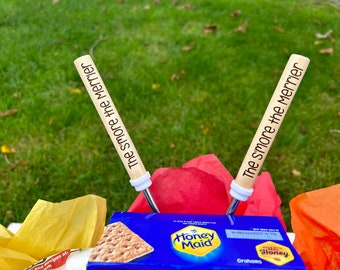 May include: Two wooden skewers with the text "The s'more the merrier" printed on them. The skewers are resting on a red, yellow, and orange tissue paper background. A box of Honey Maid graham crackers is visible in the background.