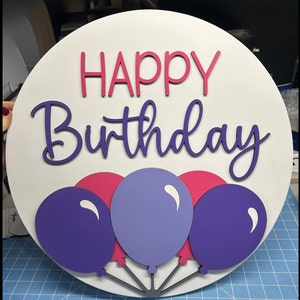 K&ouml;nnte beinhalten: Runder, wei&szlig;er Holzschild mit den Worten "HAPPY Birthday" in rosa und lila Schrift. Darunter befinden sich drei Ballons in Lila und Rosa mit grauen Schn&uuml;ren. Eine festliche Geburtstagsdekoration.