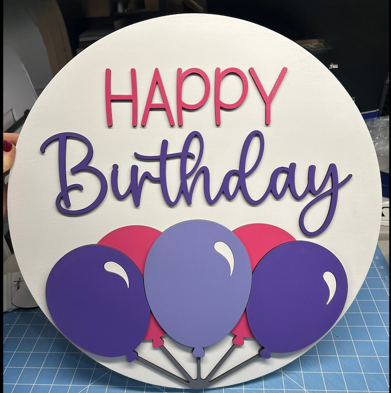 K&ouml;nnte beinhalten: Runder, wei&szlig;er Holzschild mit den Worten "HAPPY Birthday" in rosa und lila Schrift. Darunter befinden sich drei Ballons in Lila und Rosa mit grauen Schn&uuml;ren. Eine festliche Geburtstagsdekoration.