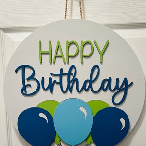 K&ouml;nnte beinhalten: Runder, wei&szlig;er Holzschild mit den Worten "HAPPY Birthday" in gr&uuml;nen und blauen Buchstaben. Darunter befinden sich drei Ballons in Blau- und Gr&uuml;nt&ouml;nen. Ein Juteseil ist zum Aufh&auml;ngen befestigt.