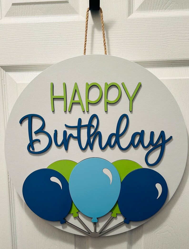 K&ouml;nnte beinhalten: Runder, wei&szlig;er Holzschild mit den Worten "HAPPY Birthday" in gr&uuml;nen und blauen Buchstaben. Darunter befinden sich drei Ballons in Blau- und Gr&uuml;nt&ouml;nen. Ein Juteseil ist zum Aufh&auml;ngen befestigt.
