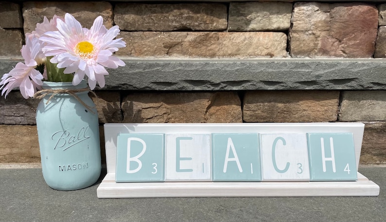 May include: A light blue mason jar with the word "Ball" and "Mason" printed on it, filled with pink flowers. The jar is sitting on a white surface in front of a wooden sign that reads "BEACH" in white letters on light blue tiles.