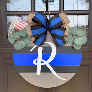 Op de afbeelding: Ronde houten deurhanger met een blauw en zwart gestreept ontwerp. Een grote witte letter "R" is gecentreerd op het hout. Een jute en blauwe strik staat bovenaan, met kunstmatige eucalyptusbladeren.