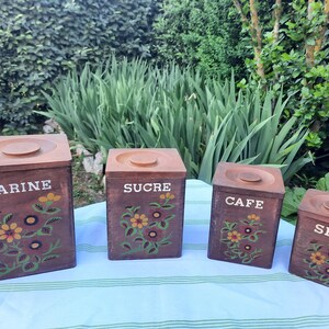 Peut inclure: Ensemble de quatre bocaux de cuisine en bois vintage. Chaque bocal marron porte l'inscription blanche : "FARINE", "SUCRE", "CAFE" et "SEL". Des motifs floraux jaunes, verts et rouges ornent le devant de chaque bocal. Les couvercles sont de couleur bois clair.