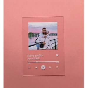 May include: A white framed digital music player with a photo of two people hugging on a bridge. The song title is "Flaw and Sins" by Juice WRLD. The music player has a play button, a heart icon, and a rewind button.