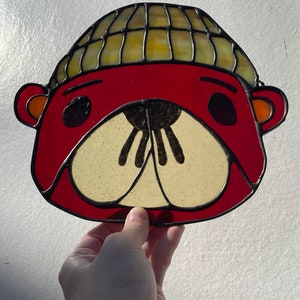 Peut inclure: Une fenêtre en verre coloré représentant un ours rouge portant un bonnet jaune et marron. L'ours a des yeux noirs et un nez noir.