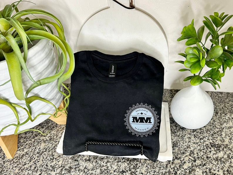 K&ouml;nnte beinhalten: Ein schwarzes T-Shirt mit einem wei&szlig;en und grauen kreisf&ouml;rmigen Design mit dem Text "MYVARA'S MM MANNER".