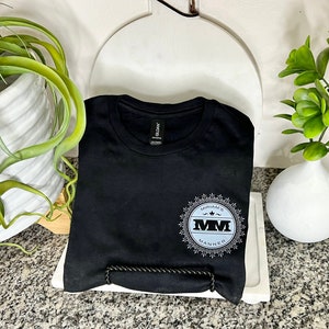 K&ouml;nnte beinhalten: Ein schwarzes T-Shirt mit einem wei&szlig;en und grauen kreisf&ouml;rmigen Design mit dem Text "MYVARA'S MM MANNER".