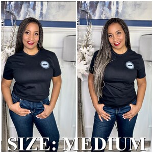 K&ouml;nnte beinhalten: Ein schwarzes T-Shirt mit einem kleinen, kreisf&ouml;rmigen Logo auf der linken Brust. Das Logo ist grau und wei&szlig; und zeigt die Buchstaben "MM" in einem Kreis. Das Model tr&auml;gt das Shirt mit einer blauen Jeans und hat graues Haar. Der Text "SIZE: MEDIUM" wird am unteren Rand des Bildes angezeigt.