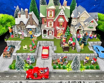 Puede incluir: Una escena de pueblo navideño en miniatura con casas, árboles, coches y personas. Las casas están decoradas con nieve y luces. Los coches son rojos, azules y marrones. Las personas llevan ropa de invierno. Hay una señal de stop en medio de la calle.