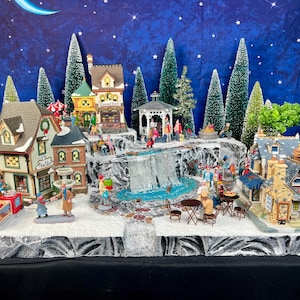 May include: A miniature Christmas village scene with houses, trees, and figures. The village features a waterfall, gazebo, and a cafe with tables and chairs. The backdrop is a starry night sky with a crescent moon.