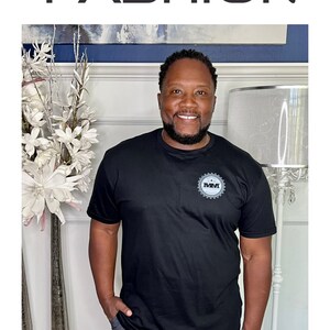 K&ouml;nnte beinhalten: Ein schwarzes T-Shirt mit einem grauen Logo, auf dem "MMI" steht. Das T-Shirt wird von einer Person mit Bart und einem L&auml;cheln getragen. Der Text "SIZE: X-LARGE" befindet sich am unteren Rand des Bildes.