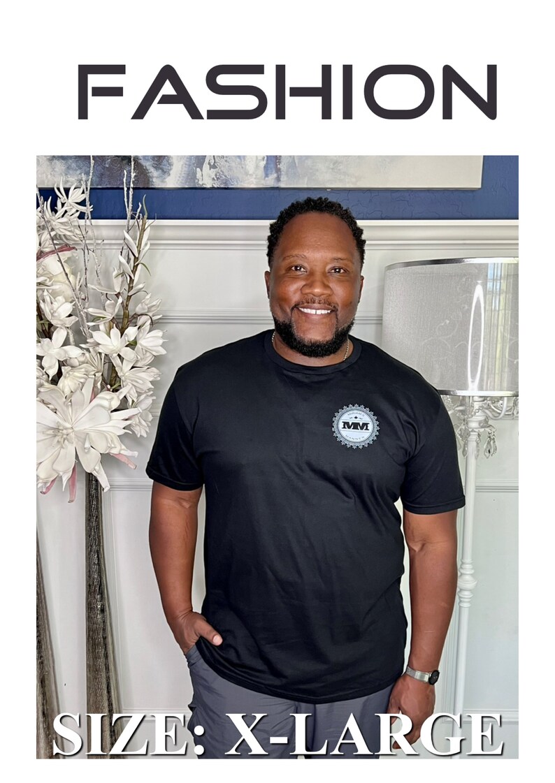 K&ouml;nnte beinhalten: Ein schwarzes T-Shirt mit einem grauen Logo, auf dem "MMI" steht. Das T-Shirt wird von einer Person mit Bart und einem L&auml;cheln getragen. Der Text "SIZE: X-LARGE" befindet sich am unteren Rand des Bildes.