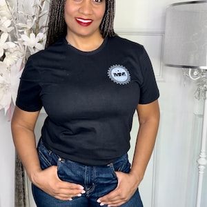K&ouml;nnte beinhalten: Eine Frau tr&auml;gt ein schwarzes T-Shirt mit einem grauen Logo, auf dem "MMVI" steht, und blaue Jeans.