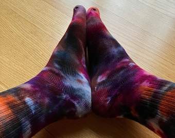 Puede incluir: Un par de calcetines coloridos con un patrón de tie-dye en tonos de morado, naranja, negro y azul. Los calcetines están doblados uno sobre el otro y están sobre un suelo de madera.