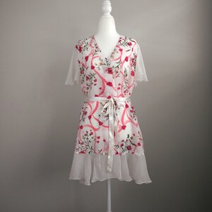 May include: A white wrap dress with a floral pattern of pink ribbons and red flowers. The dress has short sleeves, a V-neck, and a white tie belt. A ruffled hemline adds a delicate touch. The dress is displayed on a white mannequin.