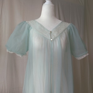 Puede incluir: Un camisón vintage azul claro con mangas cortas y transparentes. El escote y los bordes de las mangas están adornados con encaje blanco. El camisón tiene cuello en V y se abrocha con dos pequeños botones. La tela parece ser un material ligero y fluido.