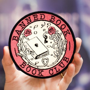 May include: A pink and black round embroidered patch with the text "Banned Book Club" and a closed book with a keyhole in the center.