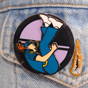May include: A black and white enamel pin featuring a cartoon character wearing a blue uniform and a yellow helmet, with red hair, upside down against a purple and black background.