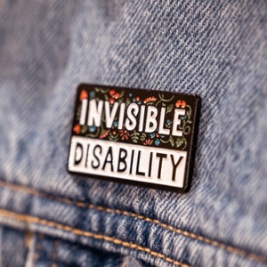 Puede incluir: Pin de esmalte negro con letras blancas que dice "Invisible Disability" rodeado de dise&ntilde;os florales coloridos. El pin est&aacute; sujeto a una chaqueta vaquera azul.