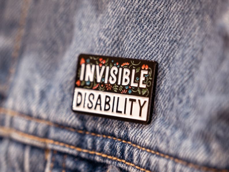 Puede incluir: Pin de esmalte negro con letras blancas que dice "Invisible Disability" rodeado de dise&ntilde;os florales coloridos. El pin est&aacute; sujeto a una chaqueta vaquera azul.