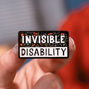Puede incluir: Un pin de esmalte negro con borde blanco y detalles florales coloridos. El pin dice "INVISIBLE DISABILITY".