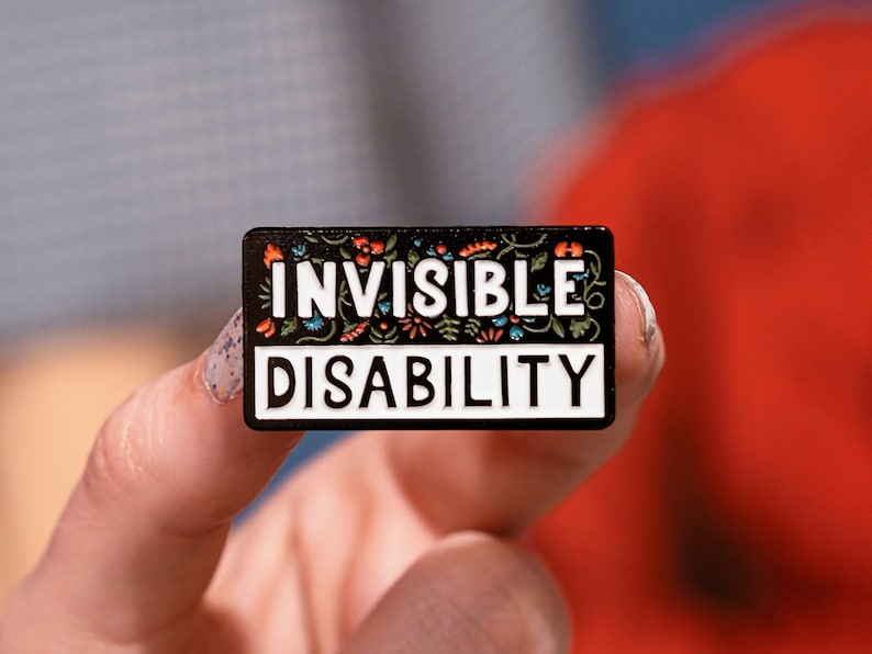 Puede incluir: Un pin de esmalte negro con borde blanco y detalles florales coloridos. El pin dice "INVISIBLE DISABILITY".