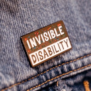 Puede incluir: Un pin de esmalte marr&oacute;n con un borde blanco y detalles florales. El pin dice "Invisible Disability" en letras blancas.