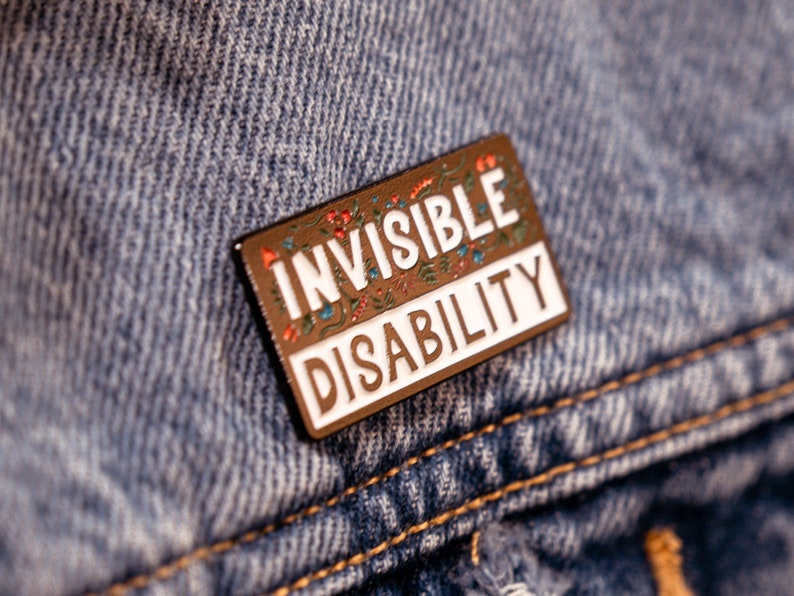 Puede incluir: Un pin de esmalte marr&oacute;n con un borde blanco y detalles florales. El pin dice "Invisible Disability" en letras blancas.