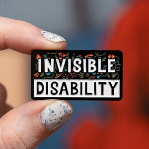 Puede incluir: Un pin de esmalte negro con letras blancas que dice "INVISIBLE DISABILITY". El pin est&aacute; decorado con dise&ntilde;os florales coloridos.