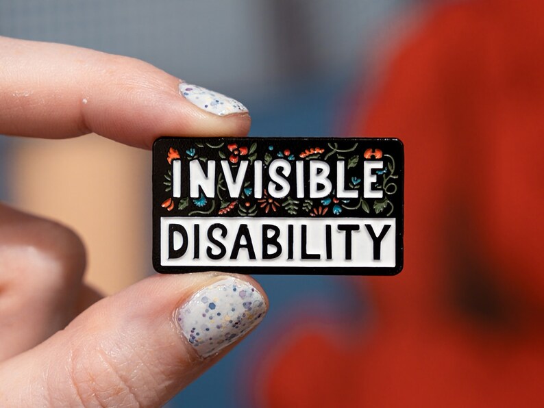 Puede incluir: Un pin de esmalte negro con letras blancas que dice "INVISIBLE DISABILITY". El pin est&aacute; decorado con dise&ntilde;os florales coloridos.