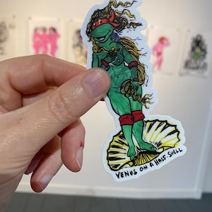 Puede incluir: Una pegatina con un personaje de dibujos animados verde, rojo y amarillo al estilo de la estatua de la "Venus de Milo". El personaje está de pie sobre una media concha con el texto "Venus on a Half-Shell" debajo.