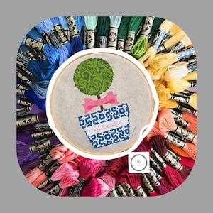 May include: A close-up of a cross-stitch embroidery hoop with a design of a green topiary in a blue and white planter with a pink ribbon. The hoop is surrounded by a variety of colorful embroidery floss.