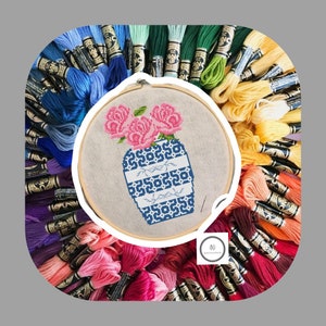 May include: A close-up of a cross-stitch embroidery project depicting a blue and white vase with pink roses. The embroidery is framed in a wooden hoop and surrounded by a variety of colorful embroidery floss.