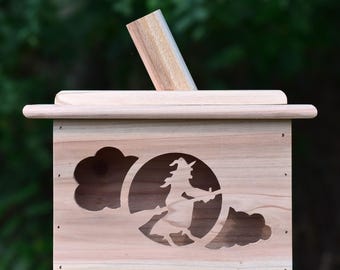 May include: A light brown wooden birdhouse featuring a witch silhouette on a broomstick cut-out. The birdhouse has a rectangular shape with a slanted roof and a wooden dowel extending from the top. Suitable for Halloween.