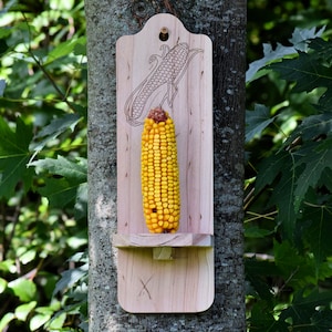 May include: A wooden bird feeder shaped like an ear of corn with a carved corncob design. The feeder has a small platform for holding corn on the cob.