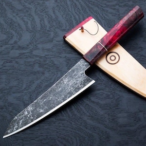 Puede incluir: Un cuchillo de chef negro y plateado con un mango de madera rojo y negro. El cuchillo está en una vaina de madera con una incrustación de madera roja y negra. La vaina tiene un círculo con una línea que lo atraviesa.