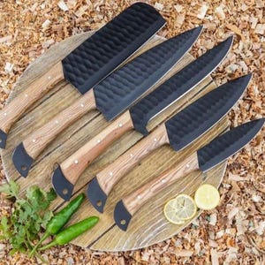 Puede incluir: Conjunto de cinco cuchillos de cocina negros con mangos de madera. Los cuchillos están dispuestos sobre una superficie de madera con virutas de madera en el fondo.