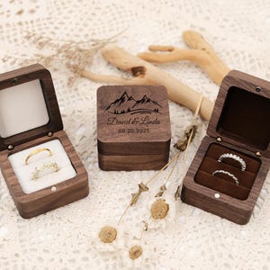Könnte beinhalten: Drei Holz-Ringboxen, zwei offen und eine geschlossen, mit Ringen. Die offenen Boxen zeigen einen goldenen Ring und einen Diamantring, während die geschlossene Box mit einer Berglandschaft und den Namen "David & Linda" und dem Datum "09.20.2025" graviert ist.