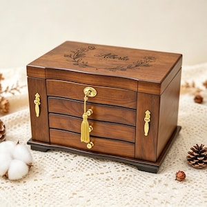 Peut inclure: Une boîte à bijoux rectangulaire en bois avec une finition brun foncé, comprenant trois tiroirs horizontaux et un couvercle supérieur. Le couvercle est gravé du nom Alberta et d'une couronne florale. Elle possède des ferrures dorées et une clé.