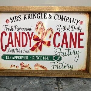 May include: A rectangular wooden sign with a vintage design. The sign features the text "MRS. KRINGLE & COMPANY" and "CANDY CANE FACTORY". Candy canes and a red bow are illustrated on the sign. The sign reads "ELF APPROVED SINCE 1847".