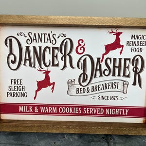 May include: Wooden framed sign with the words "Santa's Dancer & Dasher" in a vintage style. The sign includes red reindeer illustrations and text that reads "Bed & Breakfast Since 1675" and "Milk & Warm Cookies Served Nightly."