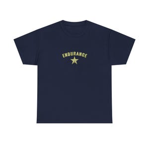 May include: A navy blue t-shirt featuring the word "ENDURANCE" in a curved font above a gold star. The design is centered on the chest. The t-shirt has a crew neck and short sleeves.