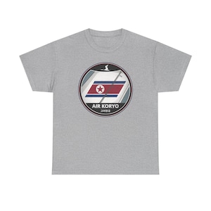 May include: A heather gray t-shirt featuring a circular graphic with the text "AIR KORYO" and Korean characters. The graphic includes a stylized flag design with a red star and a bird silhouette.